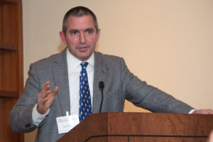 A Caucasian man with brown hair in a crew cut stands at a lectern. He's wearing a gray suit, white shirt and blue tie with white dots. He has one hand on the lectern and the other in the air waving. 