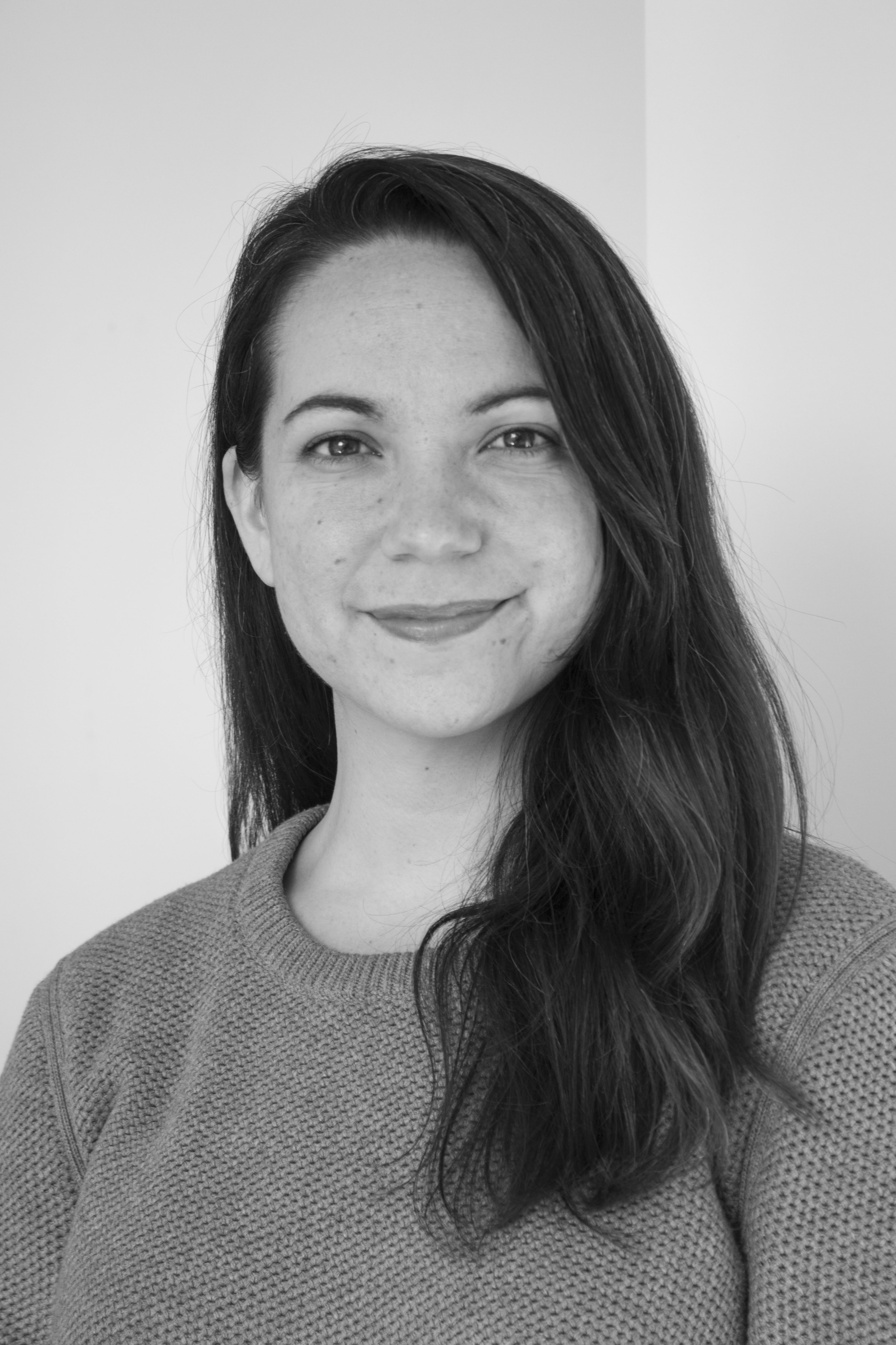 a young Caucasian woman with brown hair, parted on the right side, hanging down below her clavicle. She wears a crewneck sweater. She's smiling, but not with her teeth. 