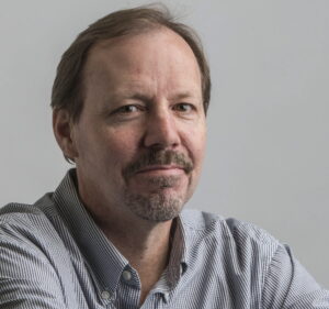 A Caucasian male with a goatee and thining brown hair, parted on the left side. He wears a checked Oxford collar shirt open at the neck. His eyes are brown and he is smiling slightly.