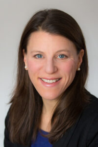 A Caucasian woman with blue eyes and brown hair down that hangs down below her collar-bone smiles.