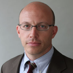 a Caucasian male looks straight at the camera. He has blue eyes and wire rime glasses. He is clean-shaven. He is mostly bald. He wears a suit, a blue Oxford collar shirt and a tie.