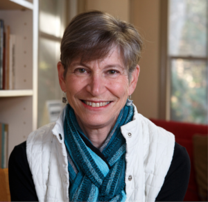 A Caucasian female with short graying brown hair smiles widely as she looks at the camera. A bookshelf is to her right, and a window is behind her left shoulder. She wears a white quilted vest, unbuttoned, over a black shirt, with a striped blue scarf around her neck.