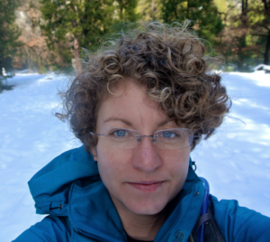 A sandy blonde Caucasian woman with blue eyes and wireless glasses stands in a snow-covered meadow.