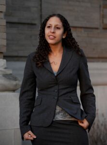 A mixed-race woman in a suitcoat and a blouse looks towards the distance. She has curly hair down below her collarbone, and is smiling.