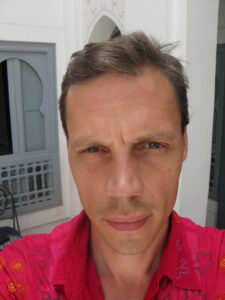 A Caucasian male with brown hair flecked with grey stares at the camera, unsmiling.