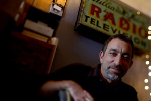 A Caucasian male with a graying goatee and short mostly brown hair smiles. He sits under a sign that looks like an old retail sign for a Radio and television store, next to a bench and cabinet.