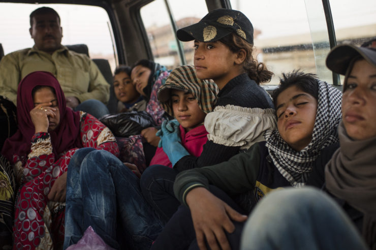 Hana, a 12-year-old Syrian refugee, is on a bus riding to work in a plum orchard in Lebanon. She hugs her cousin who is next to her. Around them are adults and young people sleeping. Hana wears a cap. Her cousin and the other women wear headscarves.