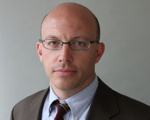 a Caucasian male looks straight at the camera. He has blue eyes and wire rime glasses. He is clean-shaven. He is mostly bald. He wears a suit, a blue Oxford collar shirt and a tie.