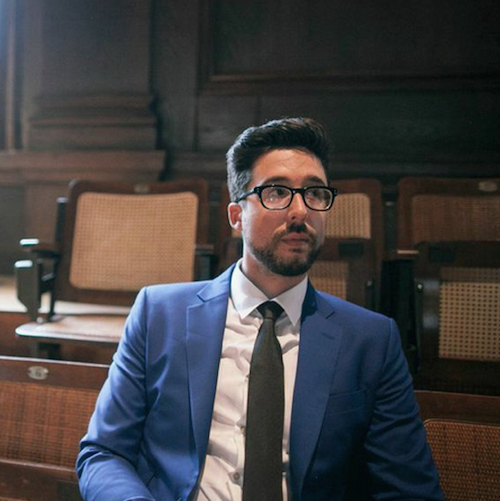 A Hispanic male with a full head of black hair and a trim beard sits in a paneled room that looks like a turn-of-the-century lecture hall. He wears black plastic-rimmed glasses, a blue sportcoat, white shirt and tie