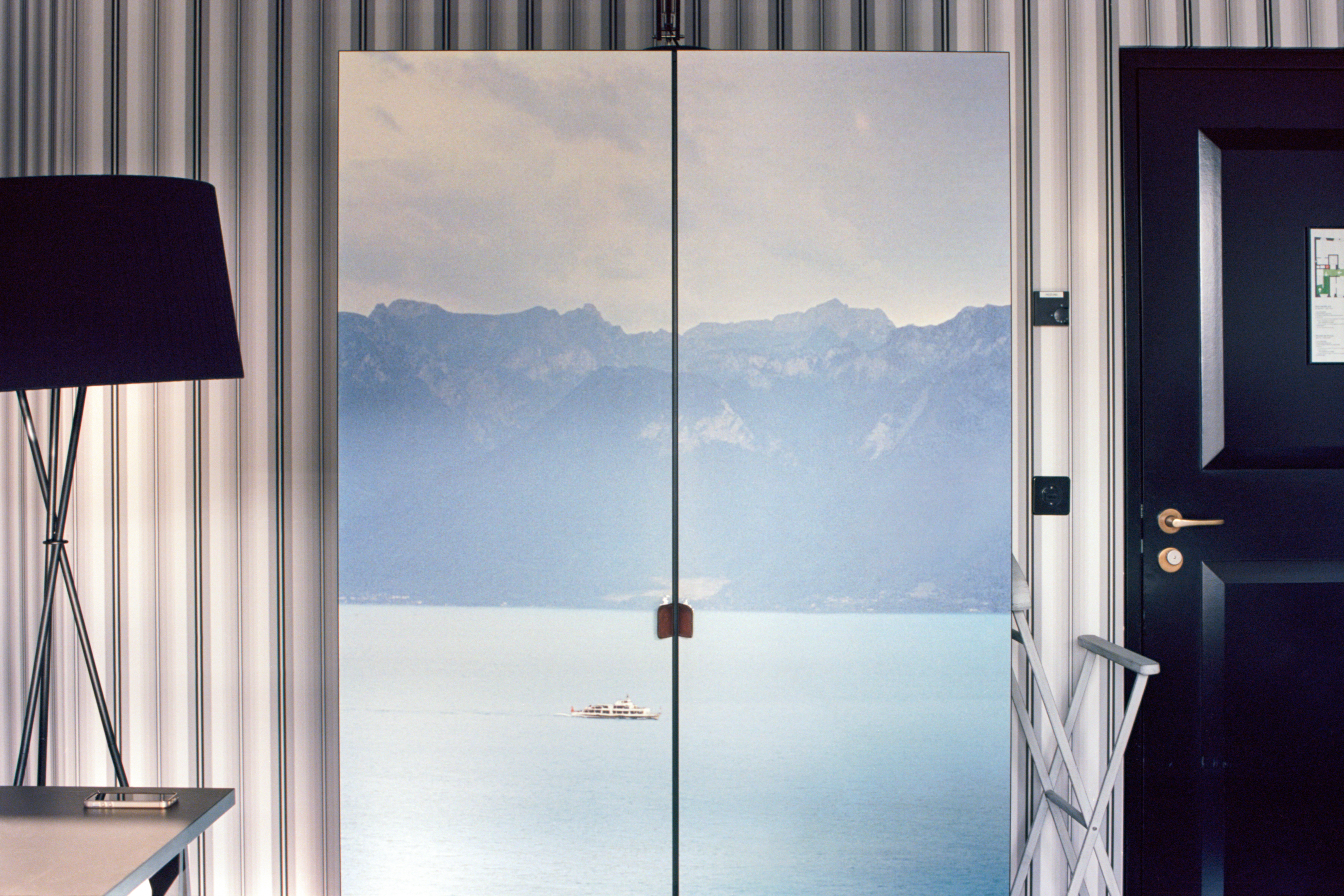 The view is what looks to be a beautiful view of the Swiss mountains behind Lake Zurich. But it is framed by a lamp and a door, and has door handles of its own. It is actually a wardrobe decorated completely by a photograph.