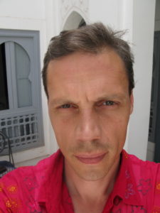 A Caucasian male with brown hair flecked with grey stares at the camera, unsmiling.
