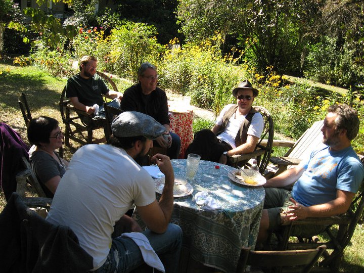 Five men and a woman sit outside around a table overlooking flowers, brush and trees. It is sunny but they are in the shade. In the center of the group sits a man in a fedora, sunglasses a white t-shirt and a vest. That is Michael Brick.