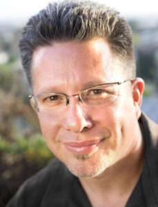 A Caucasian male with glasses, brown eyes and brown spiked hair, smiles at the camera