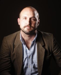 A Caucasian male, mostly bald with a brown beard and mustache, looks at the camera. He's wearing a blue denim shirt and a brown sport coat