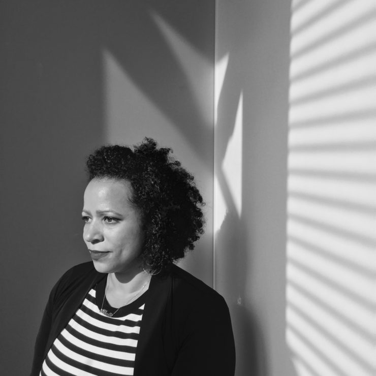 An African-American woman stands in a corner, facing out. Her hair is natural and extends to about her shoulders. She is not looking at the camera, and has a pensive expression on her face. She wears a dark jacket over a top with black and white horizontal stripes.