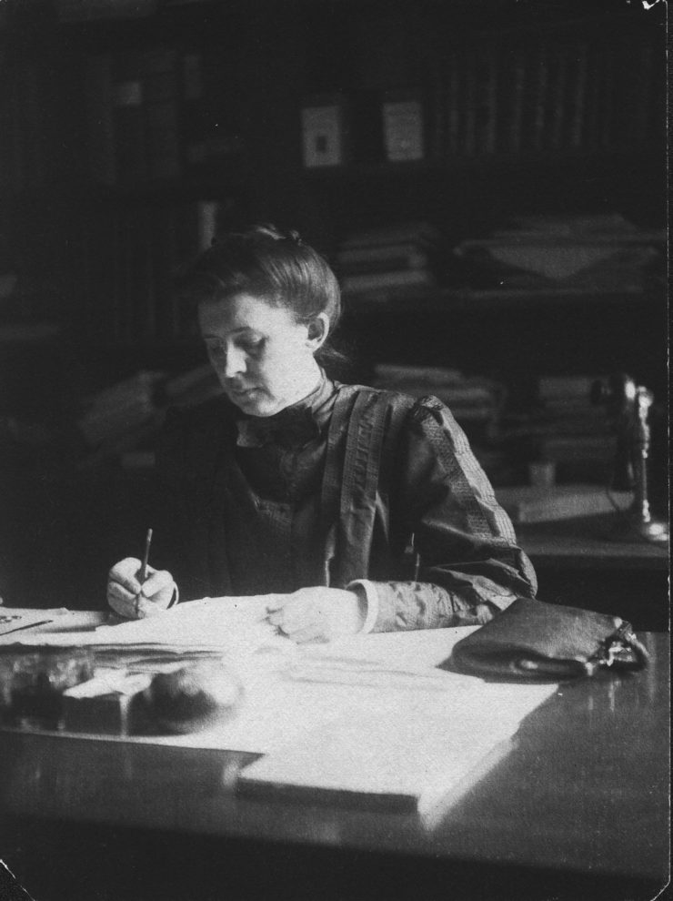 A black and white image of a woman with long hair pulled back in a bun, looking down at what she is writing. Her purse is next to her. An early telephone sits behind her.
