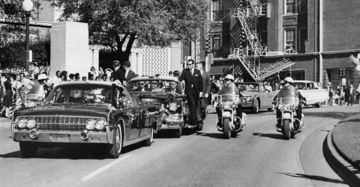 Seen through the convertible's windshield, President John F. Kennedy's hand reaches toward his head within seconds of being fatally shot. On that day, the wire car traveled several car lengths behind Kennedy’s limousine, and Merriman Smith was in the front seat.