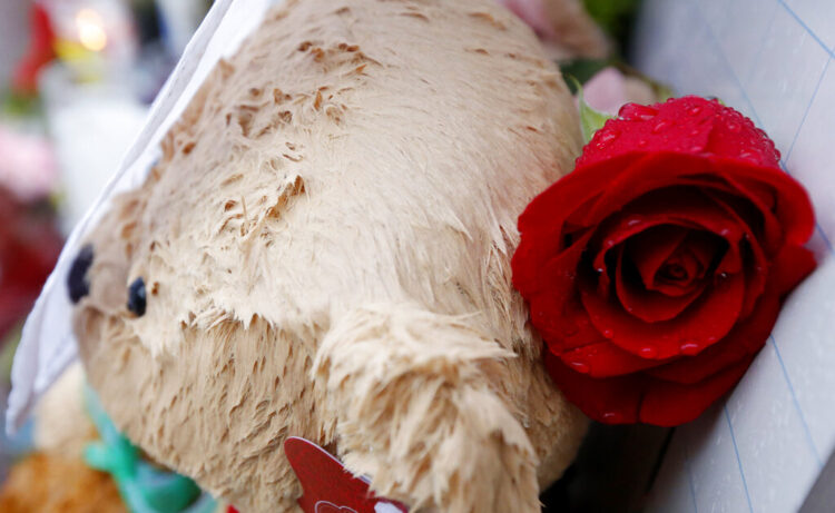 A close-up photo of a teddy bear and rose in the rain in a memorial to the victims of the 2021 mass shooting at Sandy Hook Elementary School.
